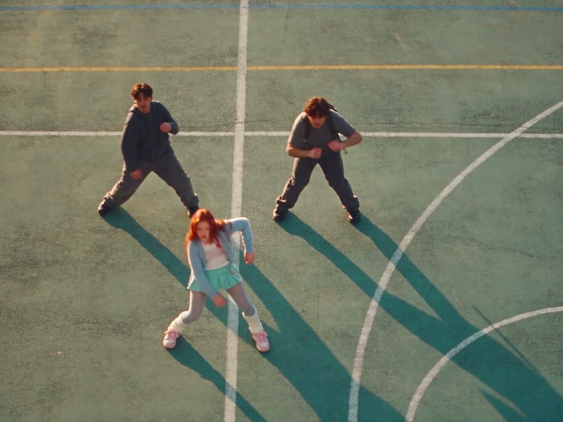 High-angle schoolyard dance on a painted court, with a synchronized crowd and a front-center lead