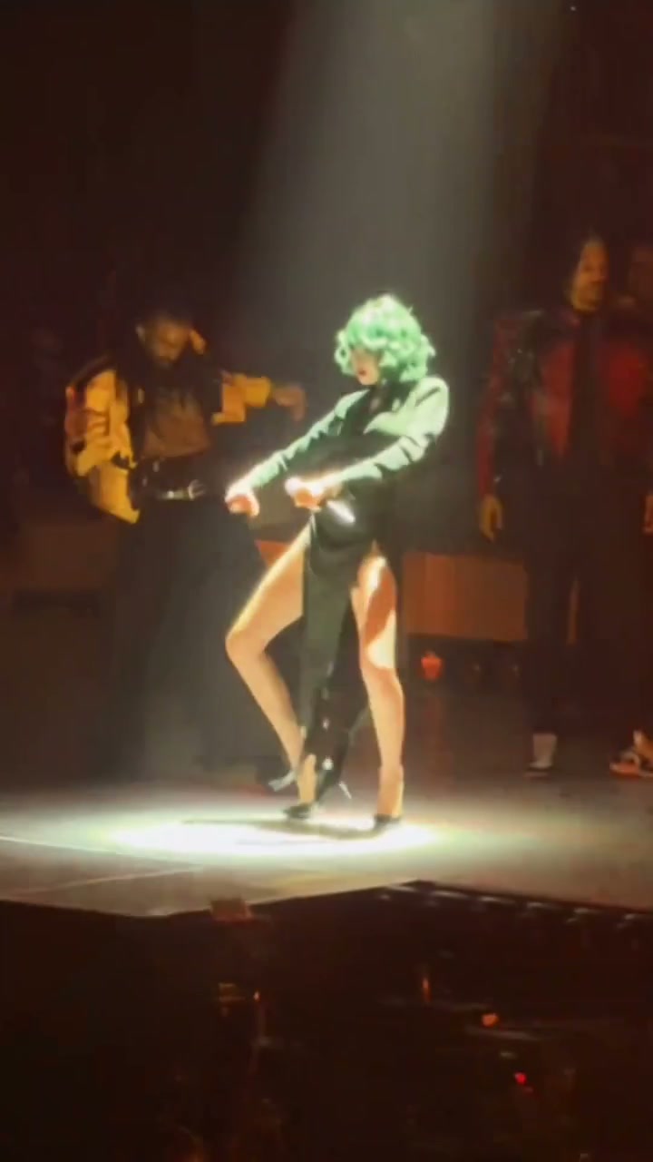 An AI-remade green-haired lead dancer in black performing with colorful-jacket backup dancers under spotlights and stage fire columns